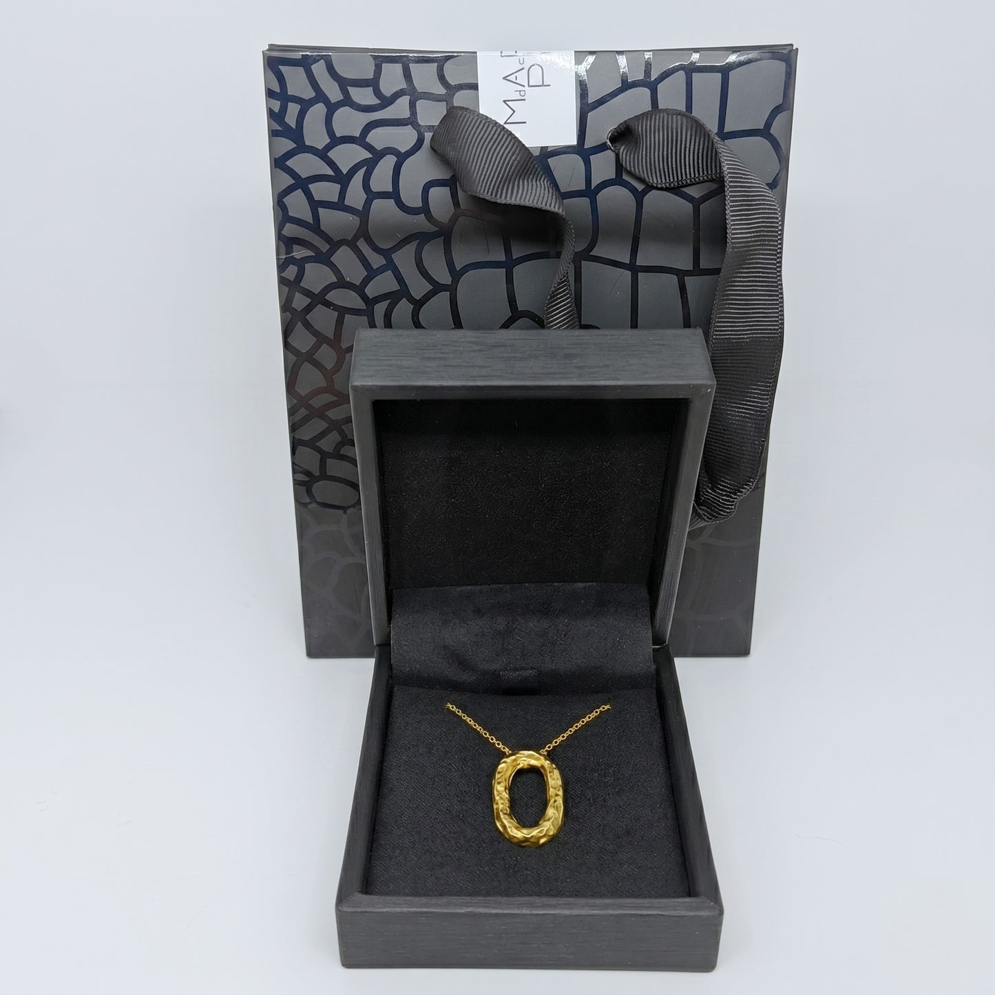 Collar ovalado de oro presentado dentro de un estuche de joyería negro abierto, con una bolsa de regalo a juego con textura de patrón geométrico al fondo.