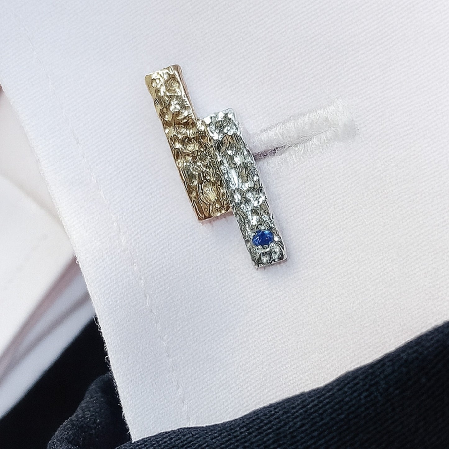 Detalle de gemelo de joyerĂa de autor colocado en el puño de una camisa blanca, mostrando la textura de plata rodiada, oro vermeil y un zafiro azul.