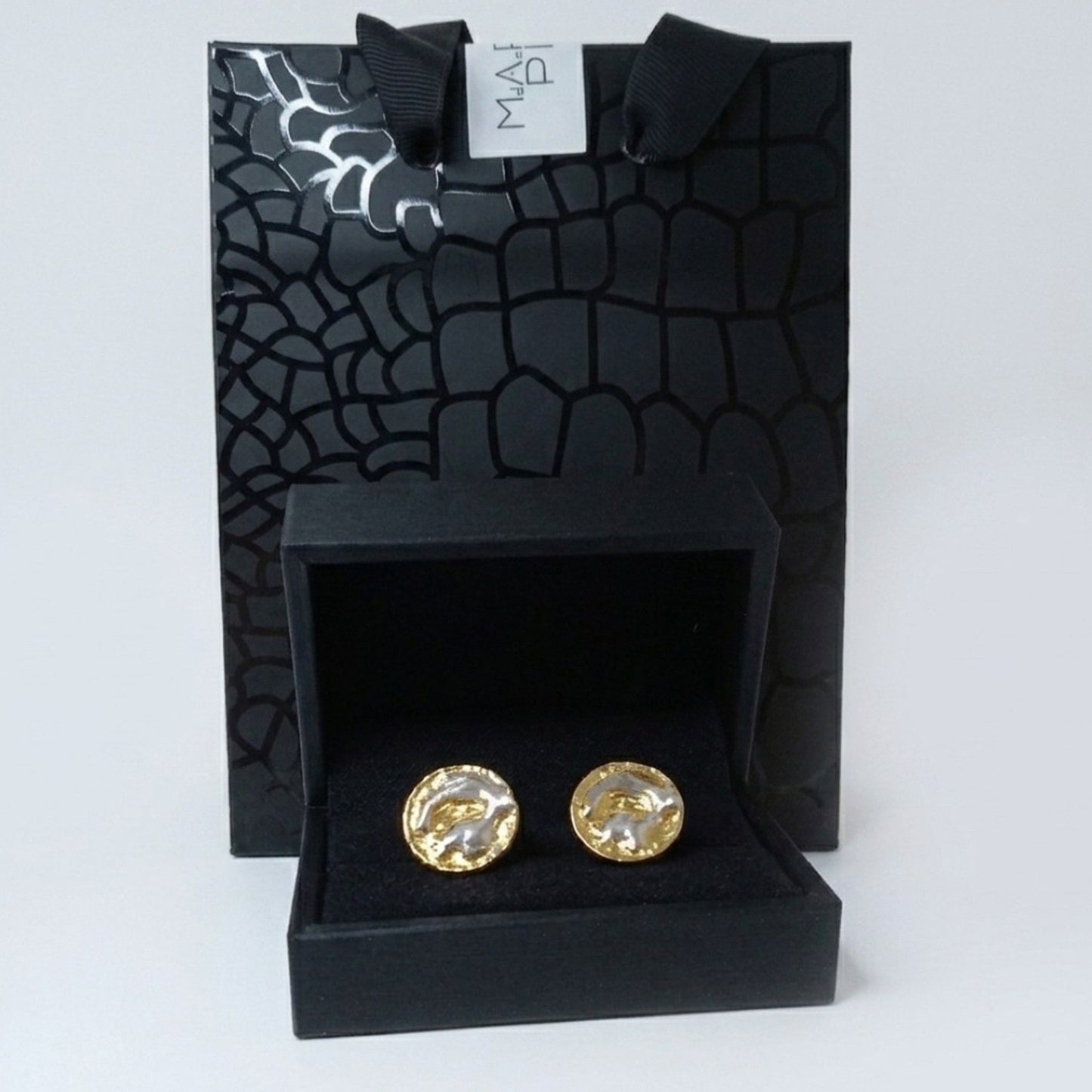 Pareja de gemelos redondos de diseño orgánico en plata y oro, presentados dentro de un estuche negro de joyería abierto. Al fondo, se muestra una bolsa de regalo negra con textura de piel de cocodrilo y una etiqueta blanca.