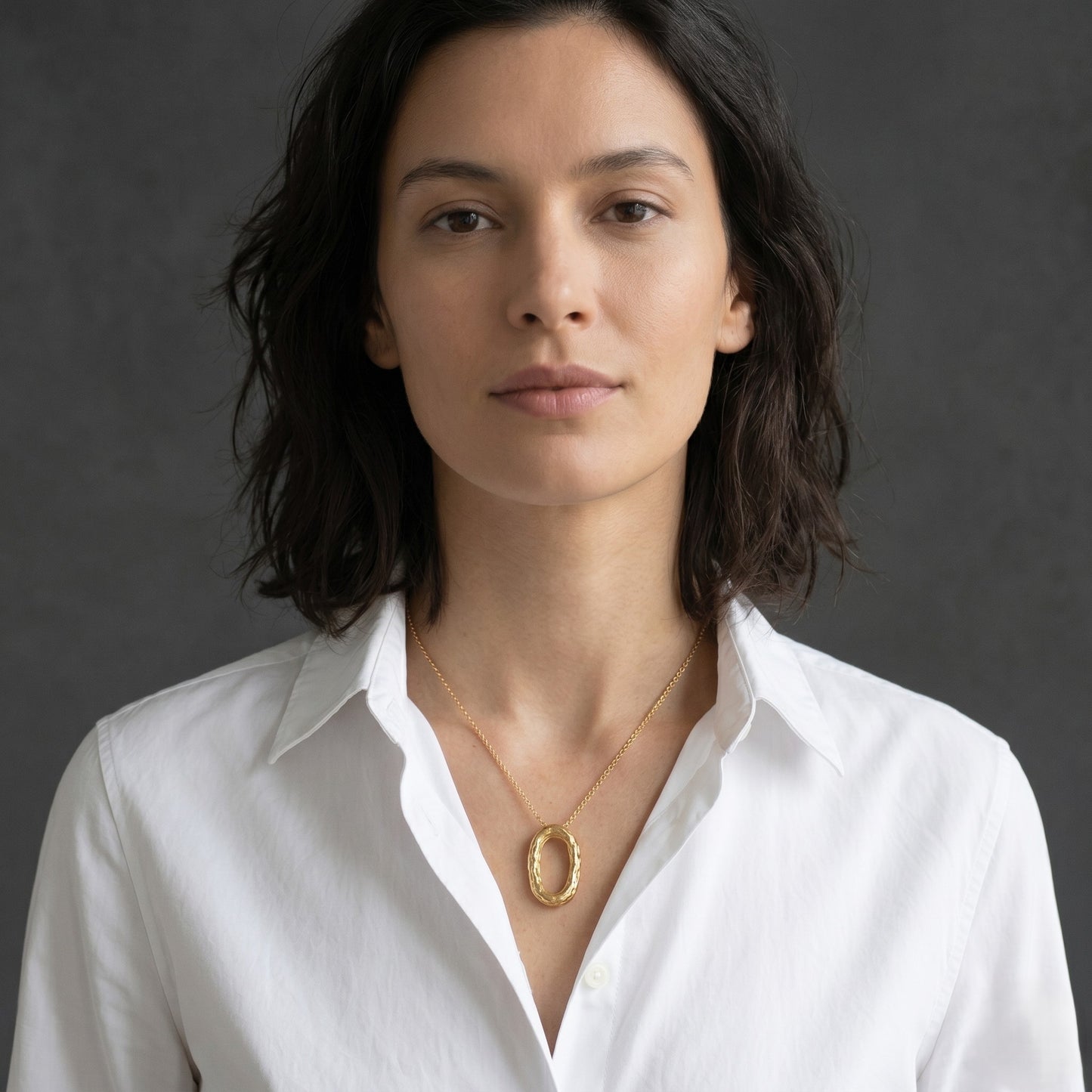 Modelo con camisa blanca luciendo el collar de cadena fina y colgante ovalado de oro texturizado, posando de frente ante un fondo gris oscuro.