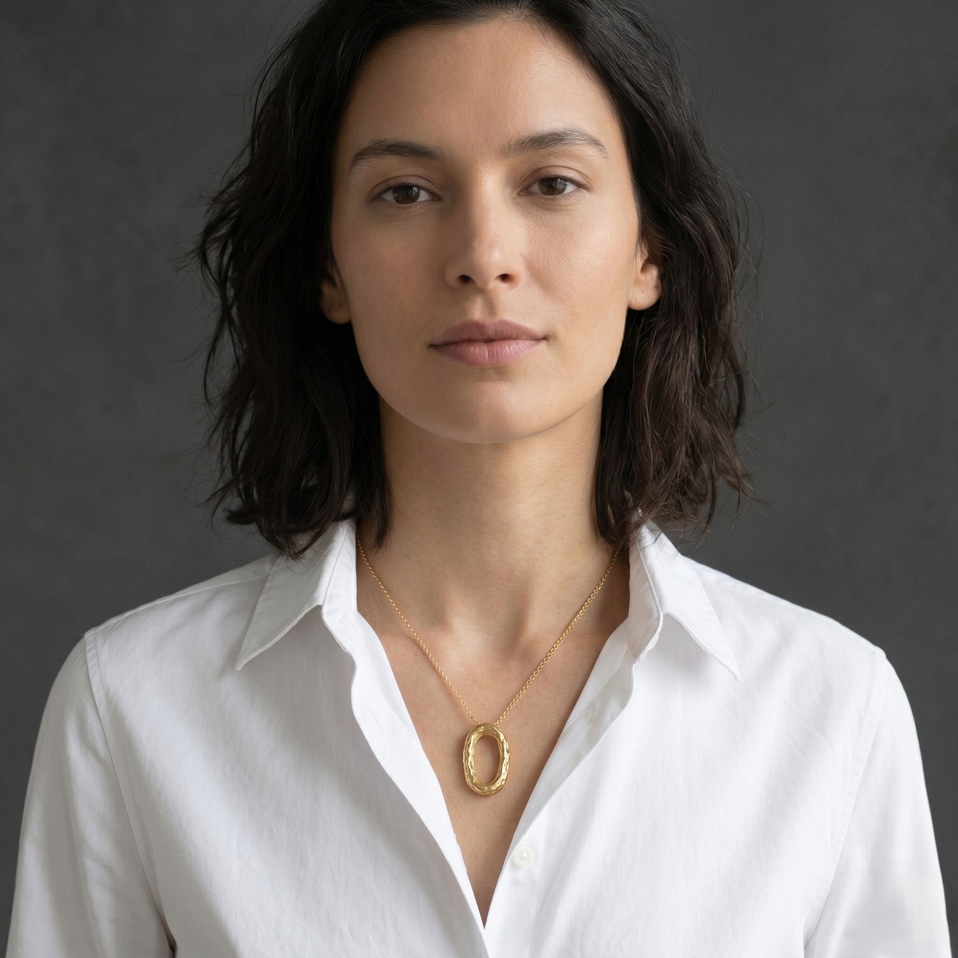 Modelo con camisa blanca luciendo el collar de cadena fina y colgante ovalado de oro texturizado, posando de frente ante un fondo gris oscuro.