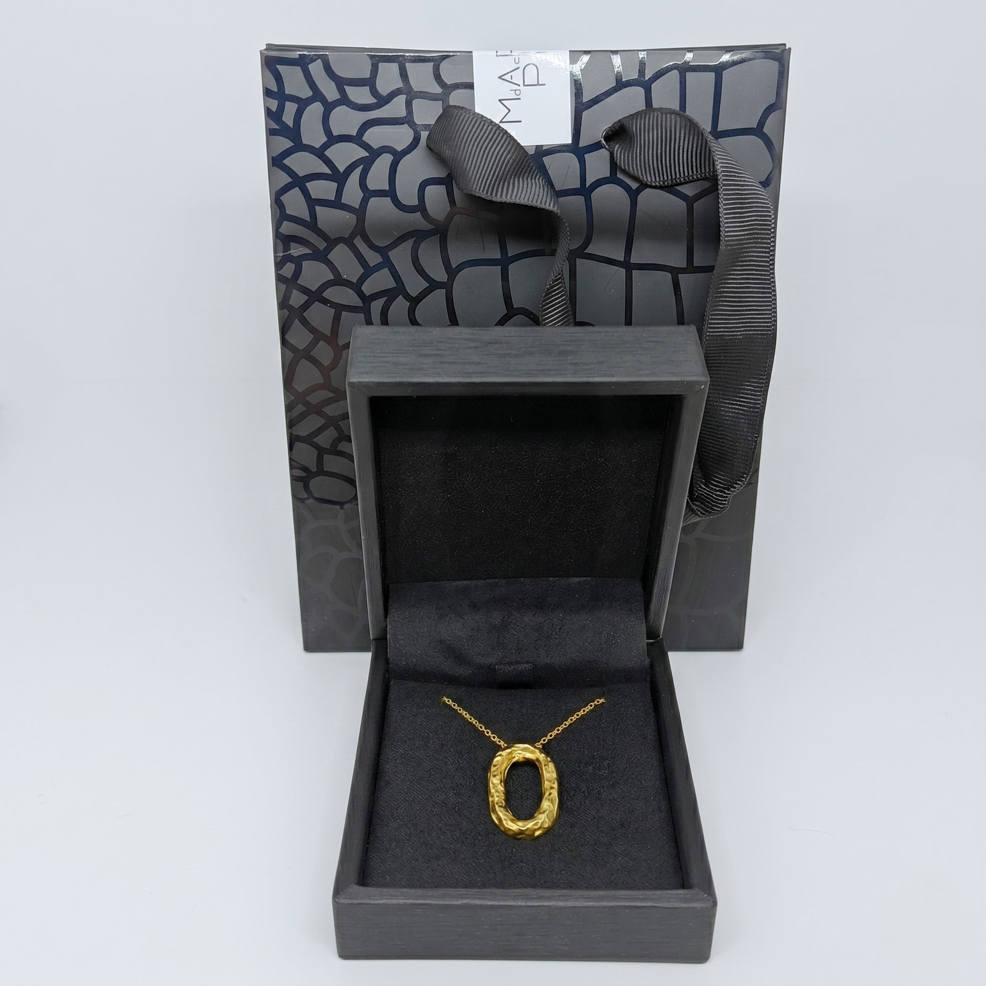 Collar ovalado de oro presentado dentro de un estuche de joyería negro abierto, con una bolsa de regalo a juego con textura de patrón geométrico al fondo.