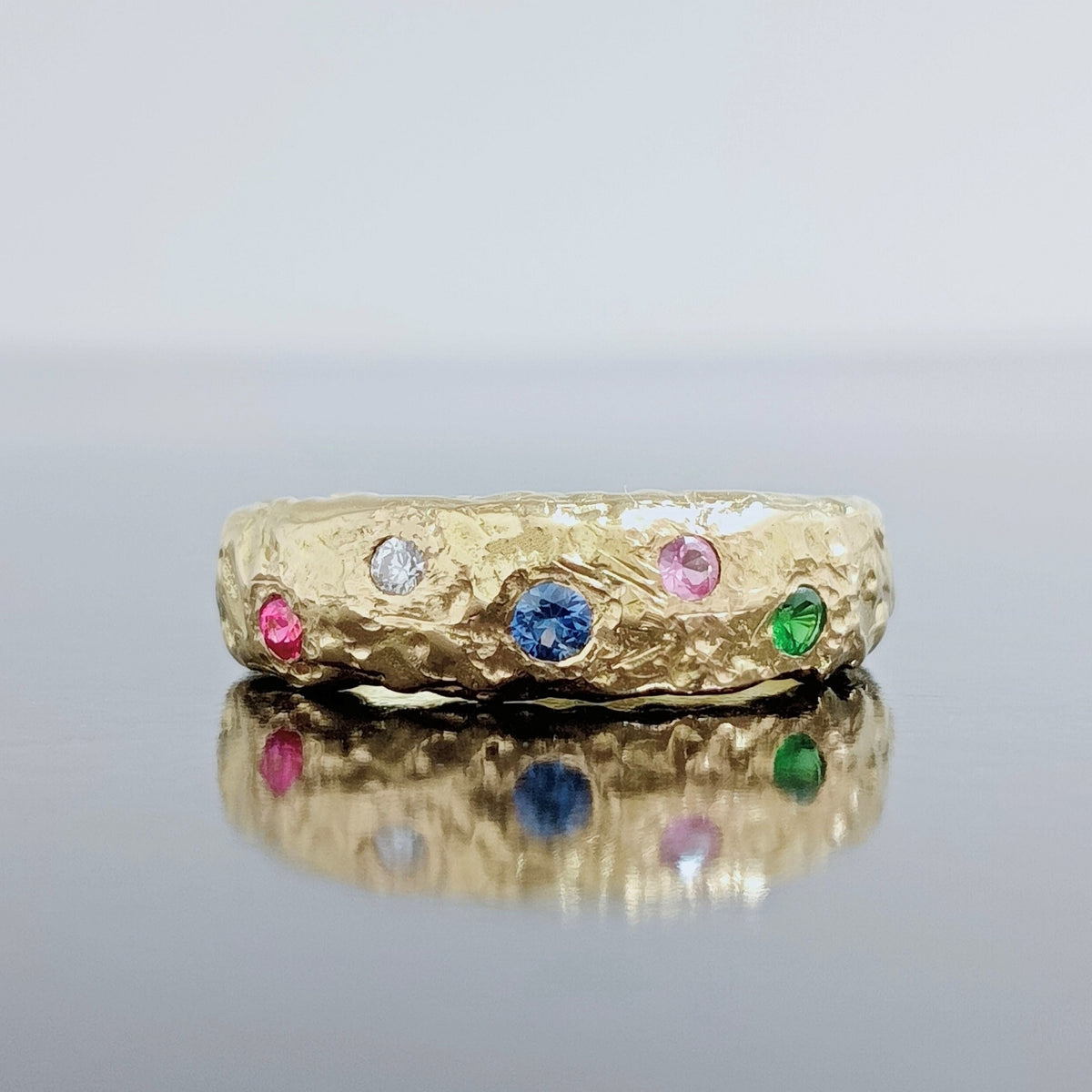 Anillo Casiopea de oro de 18 k con gemas de colores, reflejado sobre una superficie brillante