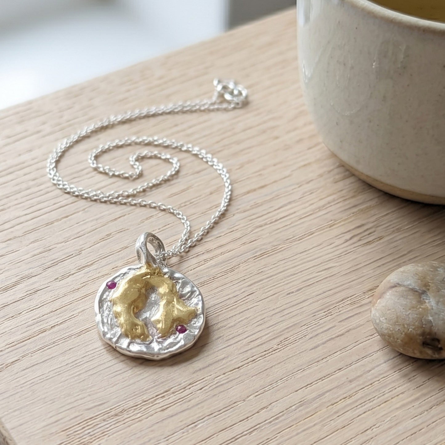 Colgante artesanal de plata y oro vermeil con dos rubíes naturales sobre mesa de madera, con cadena plateada y taza de cerámica al fondo.