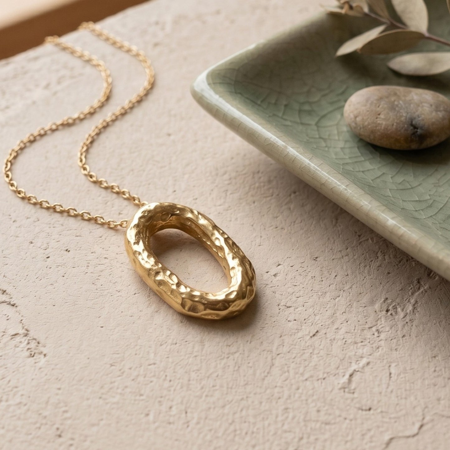 Collar de oro con colgante ovalado martillado reposando sobre una superficie beige texturizada, al lado de un plato cerámico verde con elementos decorativos naturales.