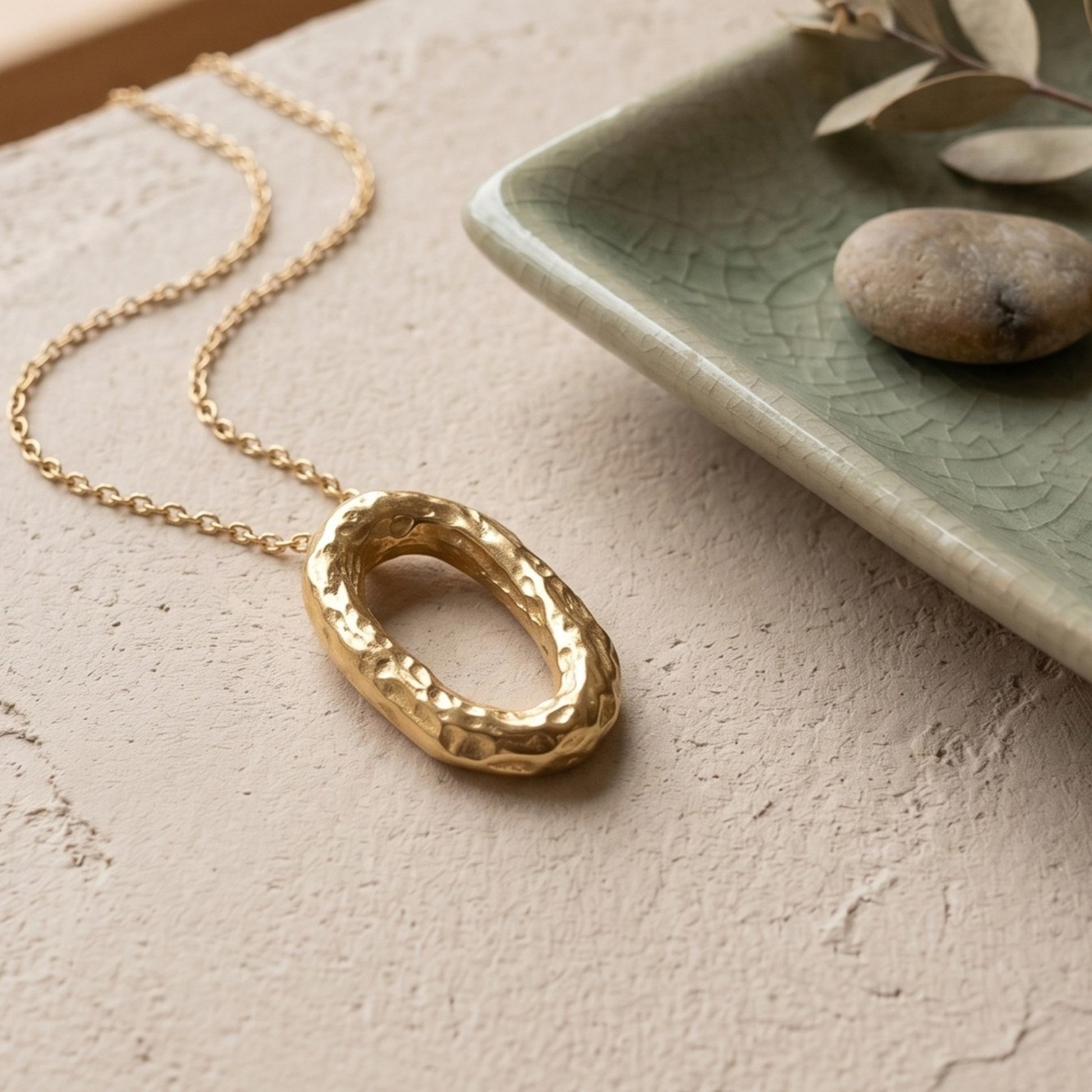 Collar de oro con colgante ovalado martillado reposando sobre una superficie beige texturizada, al lado de un plato cerámico verde con elementos decorativos naturales.