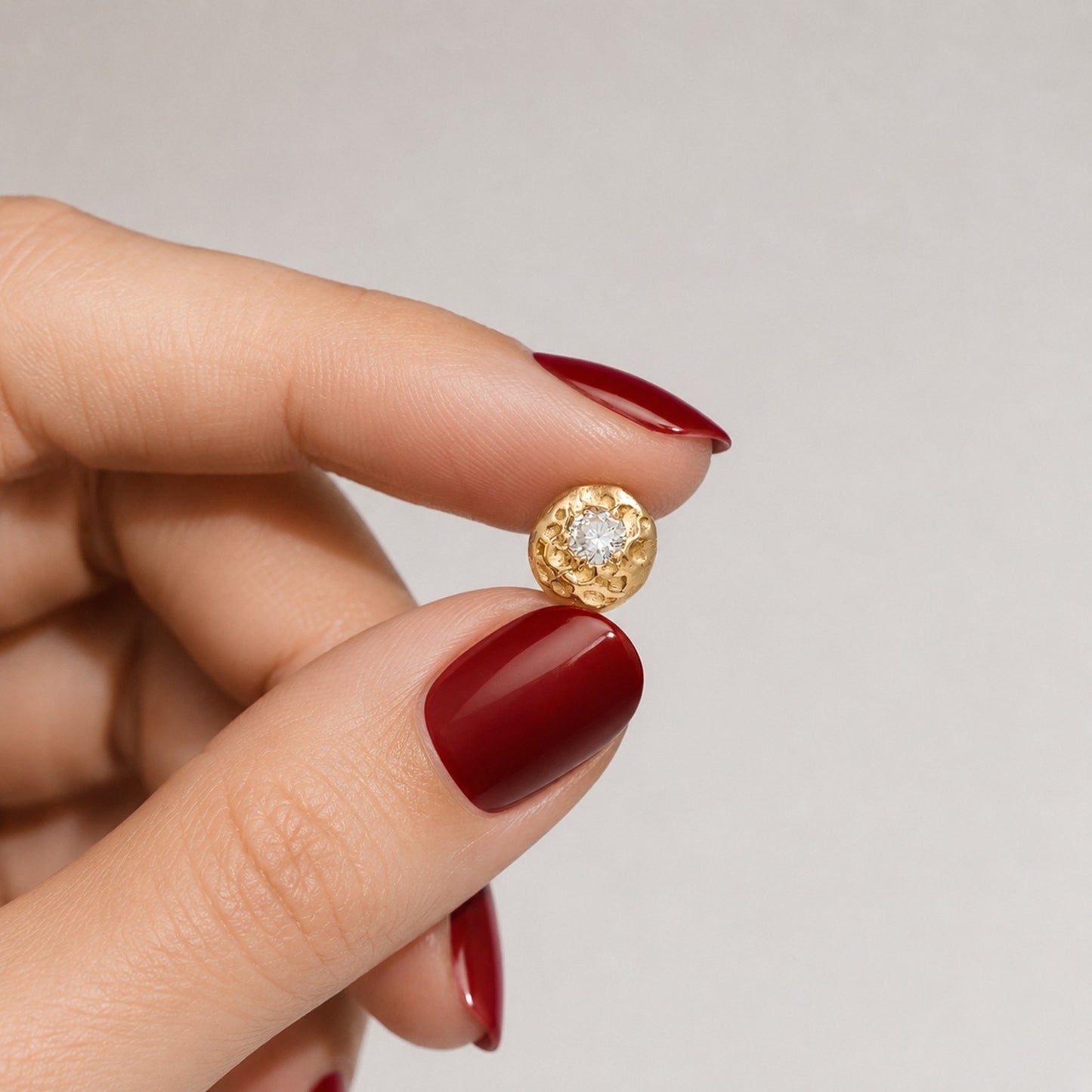 Primer plano de una mano con manicura de color rojo oscuro sosteniendo un pequeño pendiente redondo de oro texturizado para mostrar su escala real y los detalles del relieve.