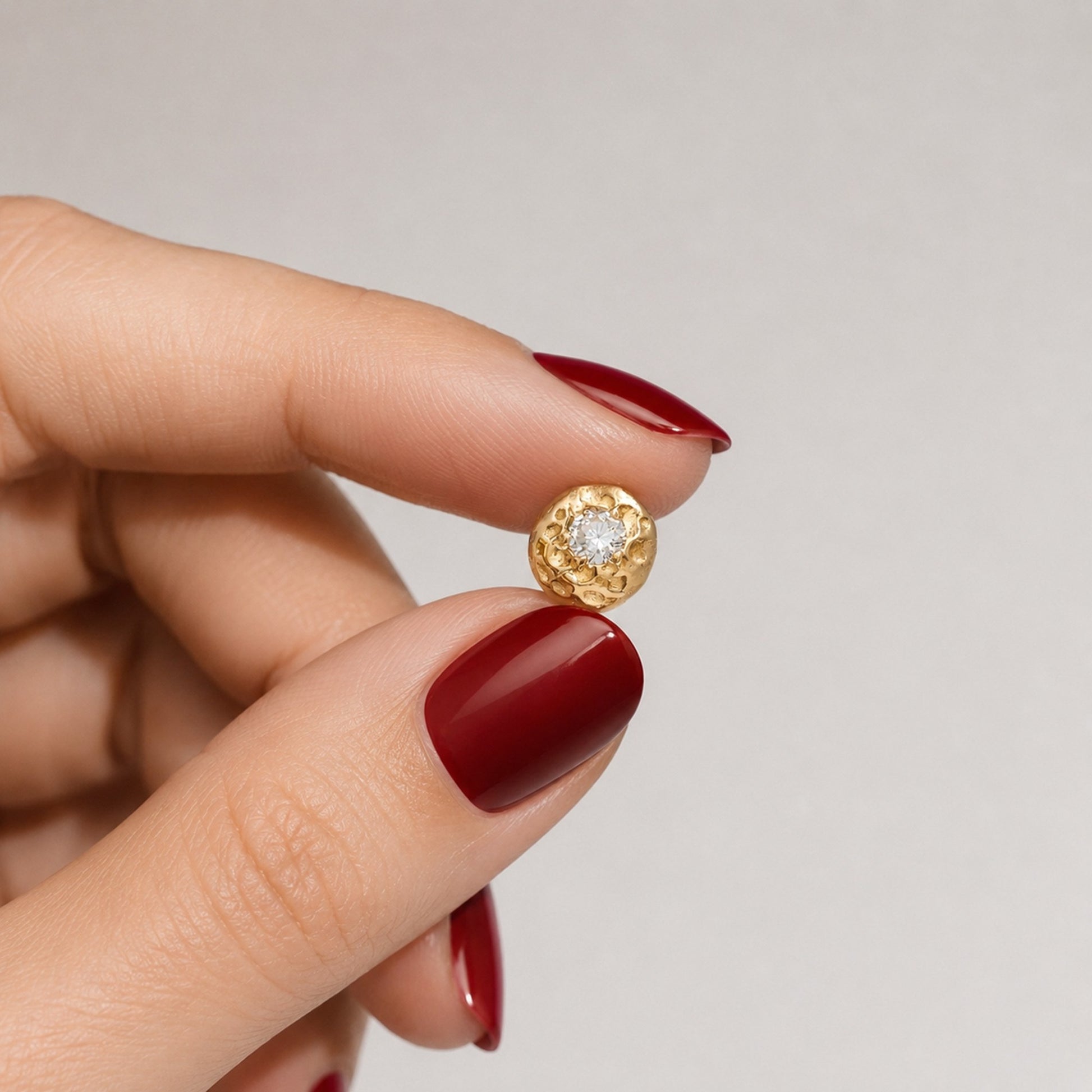 Primer plano de una mano con manicura de color rojo oscuro sosteniendo un pequeño pendiente redondo de oro texturizado para mostrar su escala real y los detalles del relieve.