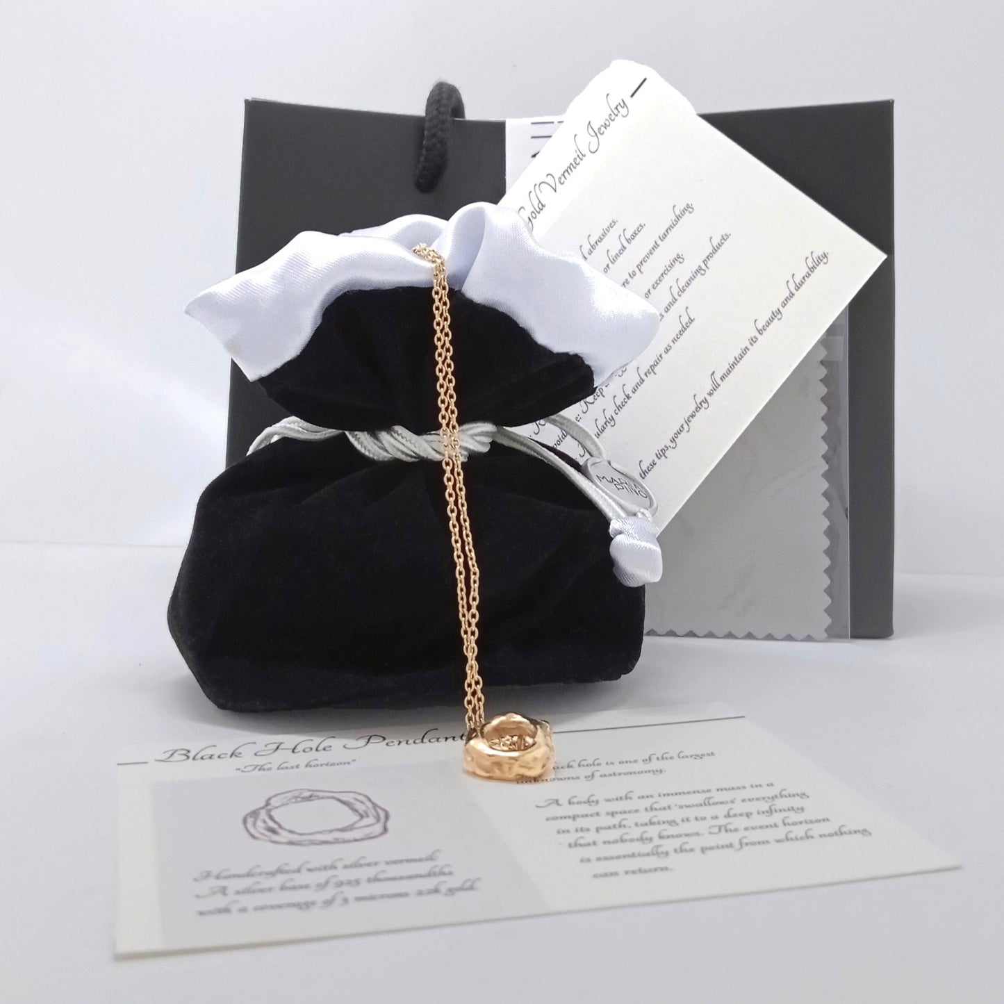 Set de regalo que incluye el colgante de oro vermeil sobre una tarjeta descriptiva "Black Hole Pendant", junto a una bolsa de terciopelo negro con bordes blancos y una caja de lujo al fondo.