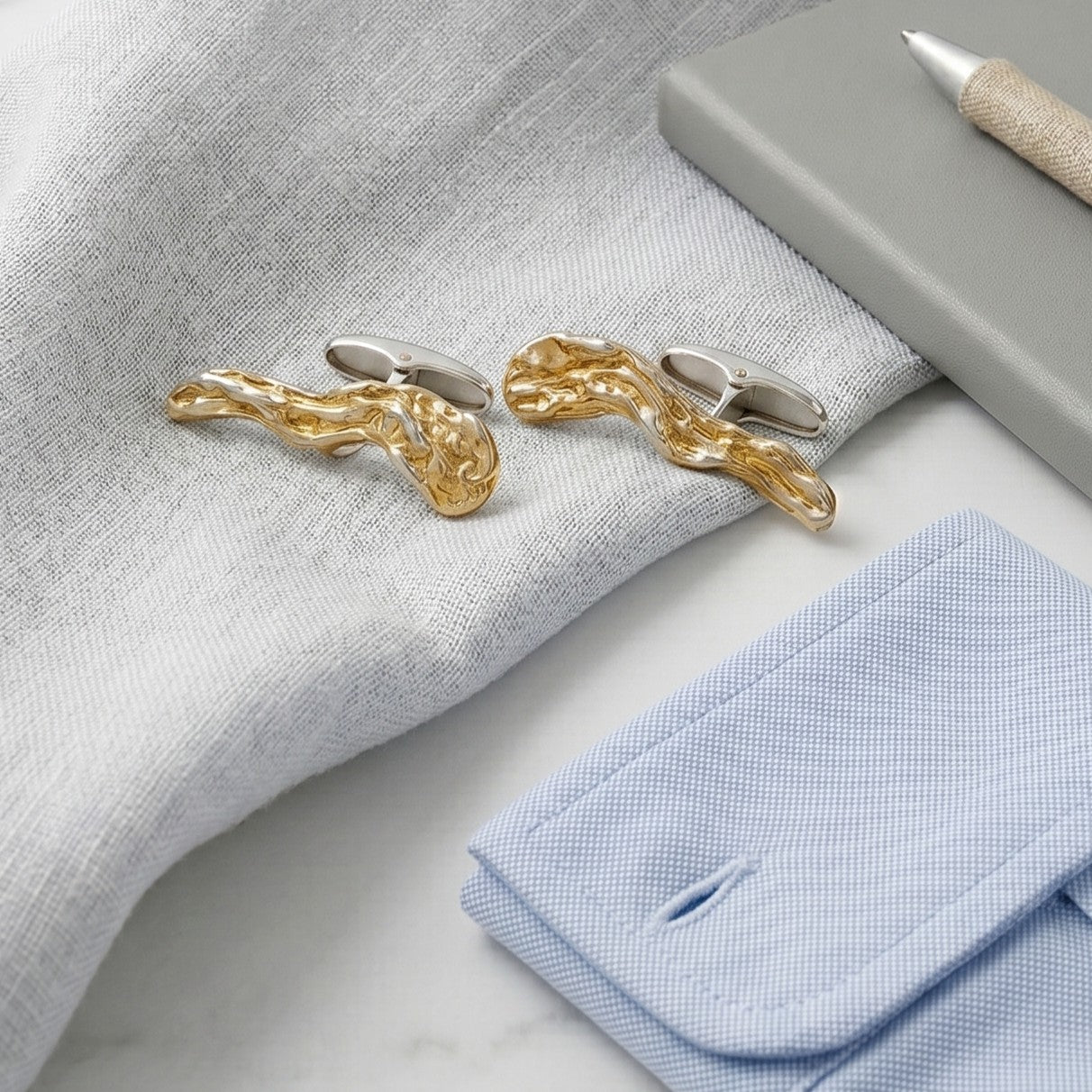 Bodegón minimalista de gemelos artesanales dorados sobre lino gris junto a puño de camisa azul claro y agenda.
