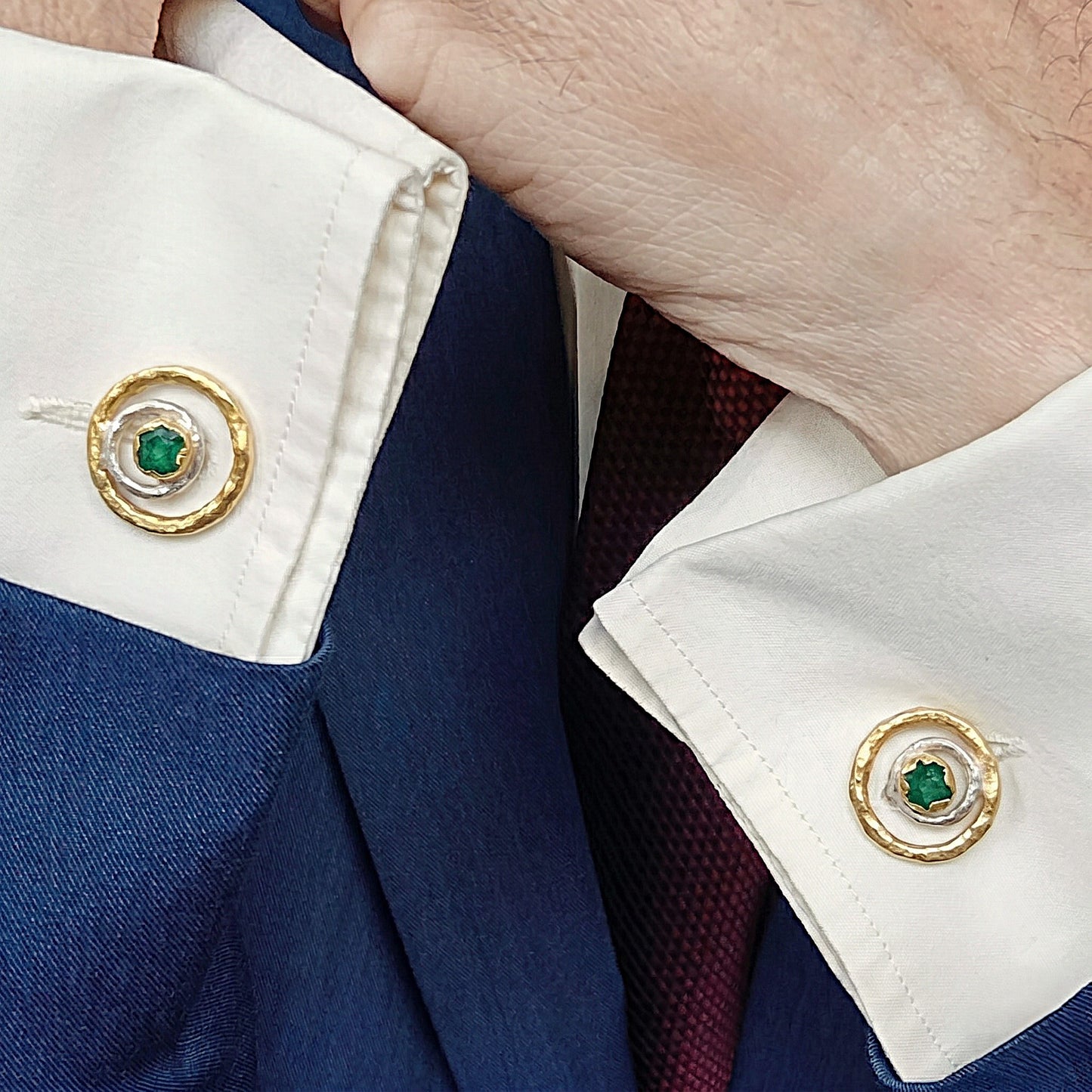 Primer plano de un hombre que lleva gemelos de esmeralda cruda en una camisa de vestir blanca, con una chaqueta azul marino y una corbata burdeos.