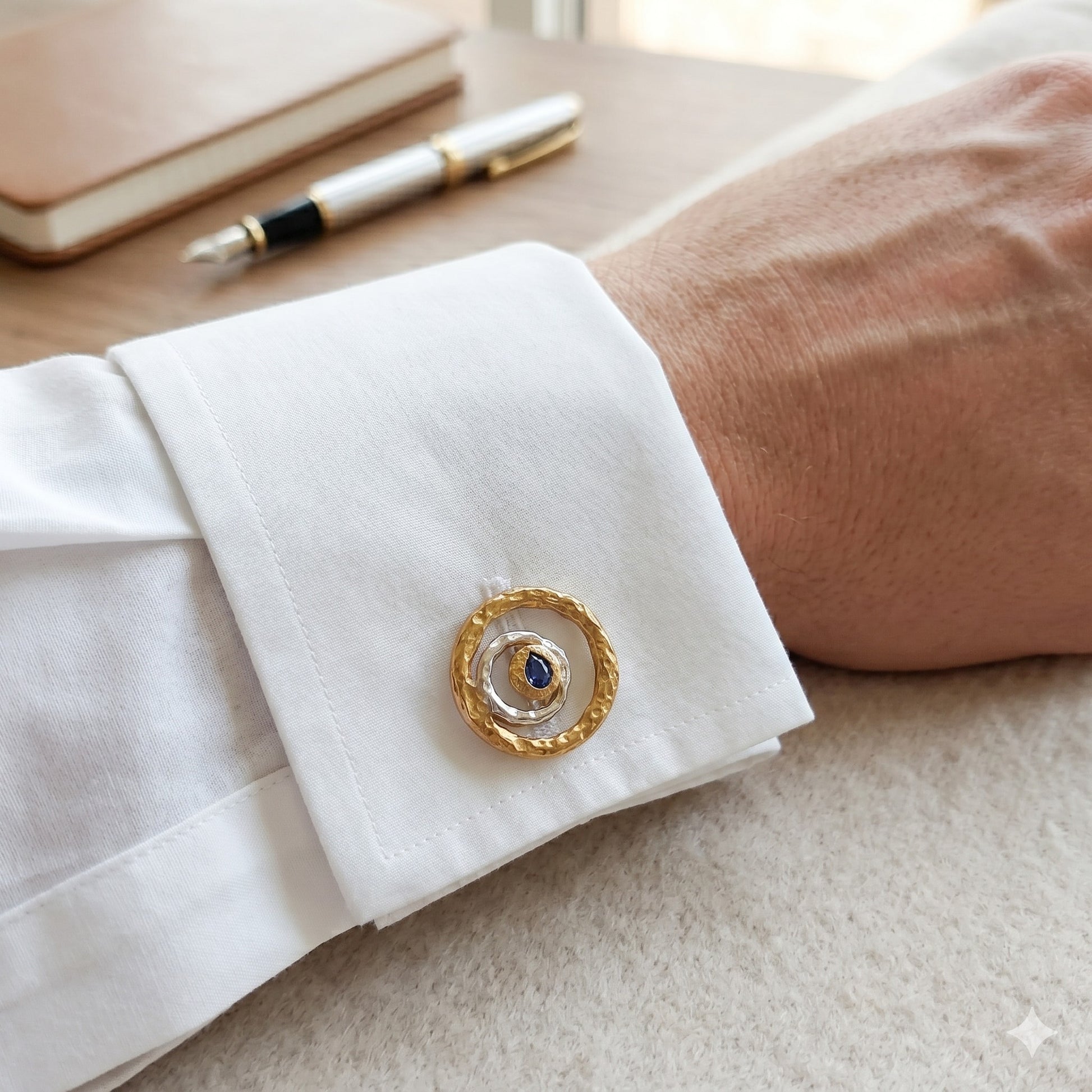 Hombre luciendo gemelos de oro y zafiro en camisa blanca de gemelos, con pluma y libreta al fondo.
