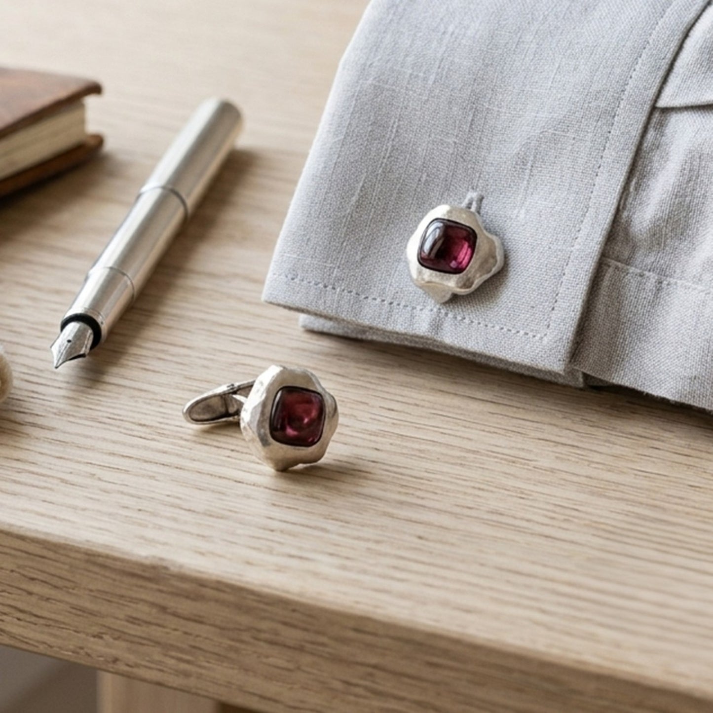 Gemelos de plata artesanal y granate junto a una pluma estilográfica sobre mesa de madera.