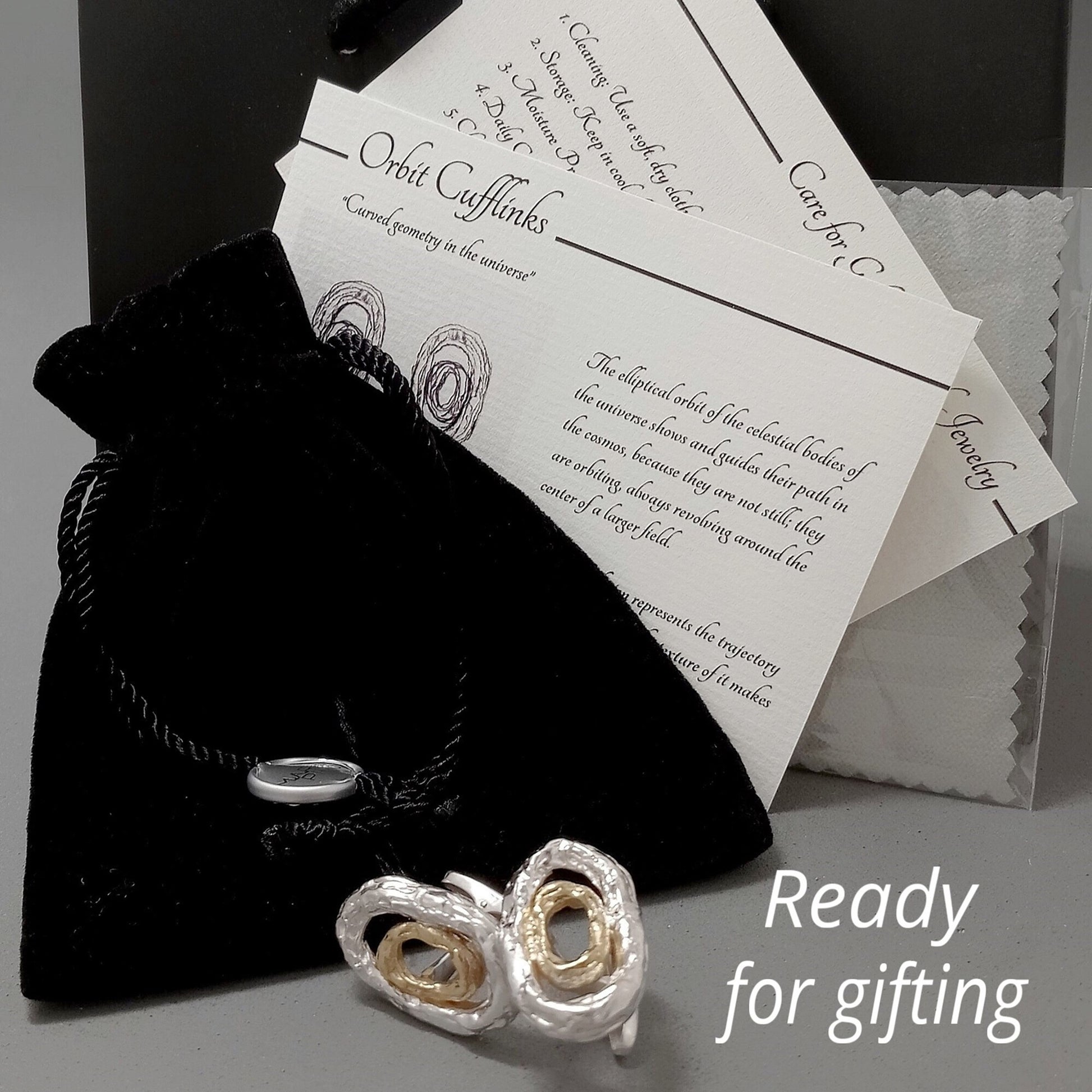 Un set de regalo para gemelos 'Órbita' que incluye una bolsa de terciopelo negro, dos tarjetas de instrucciones con diagramas de órbita concéntrica y el texto 'Care for Jewelry' y 'Orbit Cufflinks'. El par de gemelos está en primer plano sobre una superficie gris, con el texto superpuesto 'Ready for gifting'.