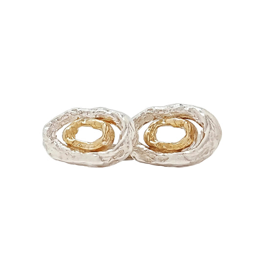 Primer plano de estudio de un par de gemelos de diseño elíptico en plata de ley texturizada con anillos interiores concéntricos de oro vermeil. Uno está de frente y el otro de perfil para mostrar el mecanismo de cierre de barra T. Fondo blanco minimalista.