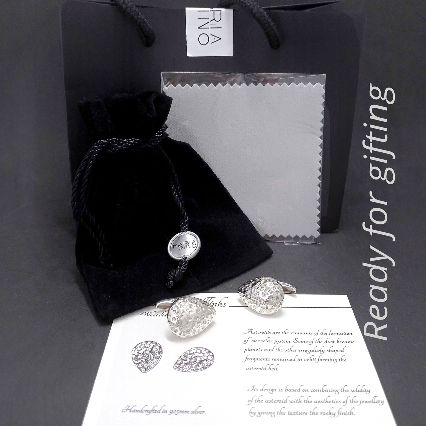 Conjunto de presentación de regalo para los gemelos de plata martillada. Incluye una bolsa de regalo de papel negro con logo, una bolsita de terciopelo negro, un paño de limpieza gris, una tarjeta con información sobre el diseño inspirado en asteroides y los propios gemelos de plata.