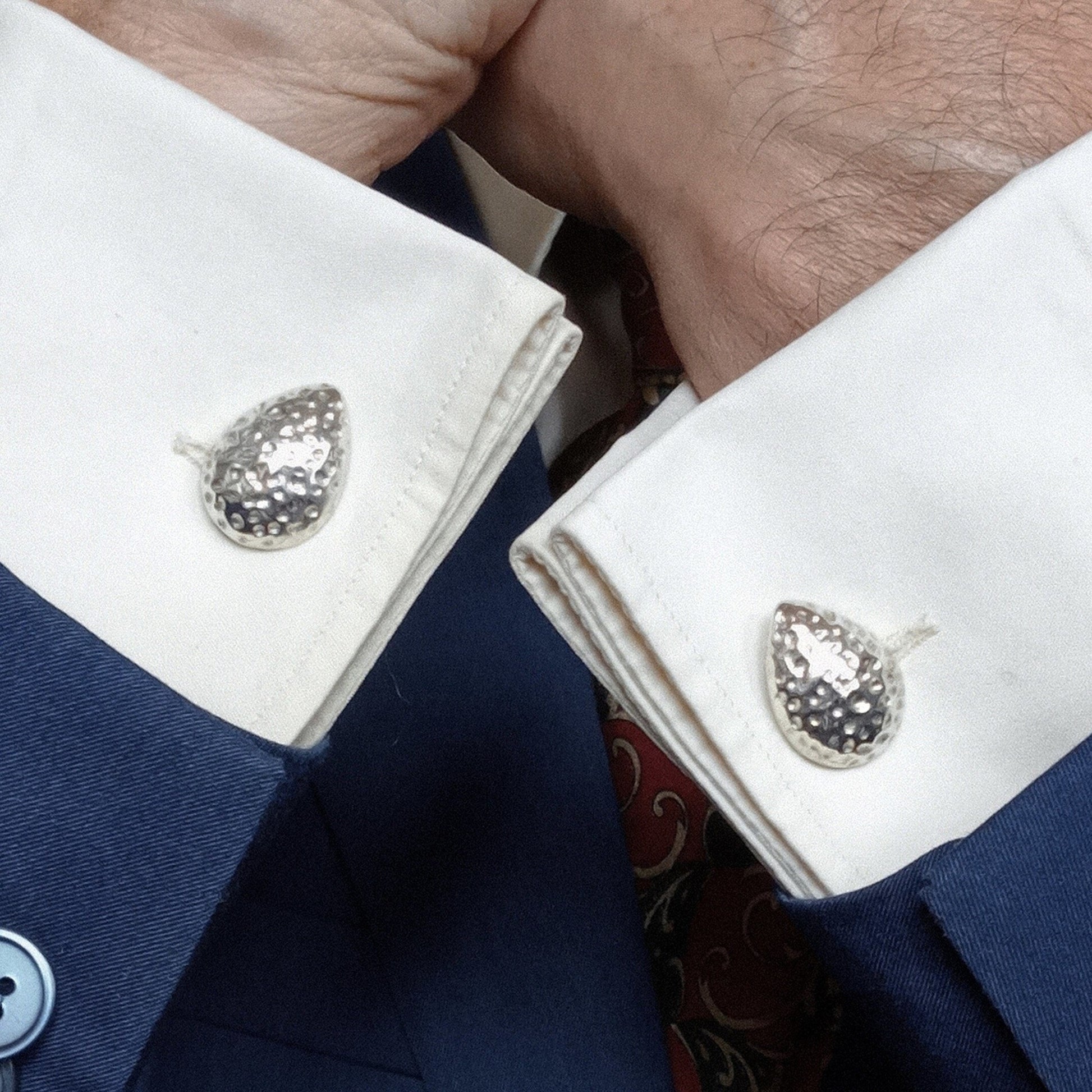 Primer plano de los gemelos de plata martillada colocados en los puños de una camisa de doble puño color marfil y una chaqueta de traje azul marino. Las manos de un hombre están entrelazadas en primer plano.