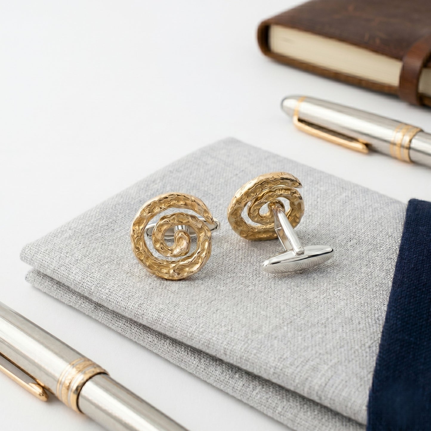 Gemelos de plata y oro vermeil con diseño espiral texturizado hecho a mano, inspirados en ondas y galaxias, presentados sobre pañuelo gris elegante junto a pluma estilográfica.