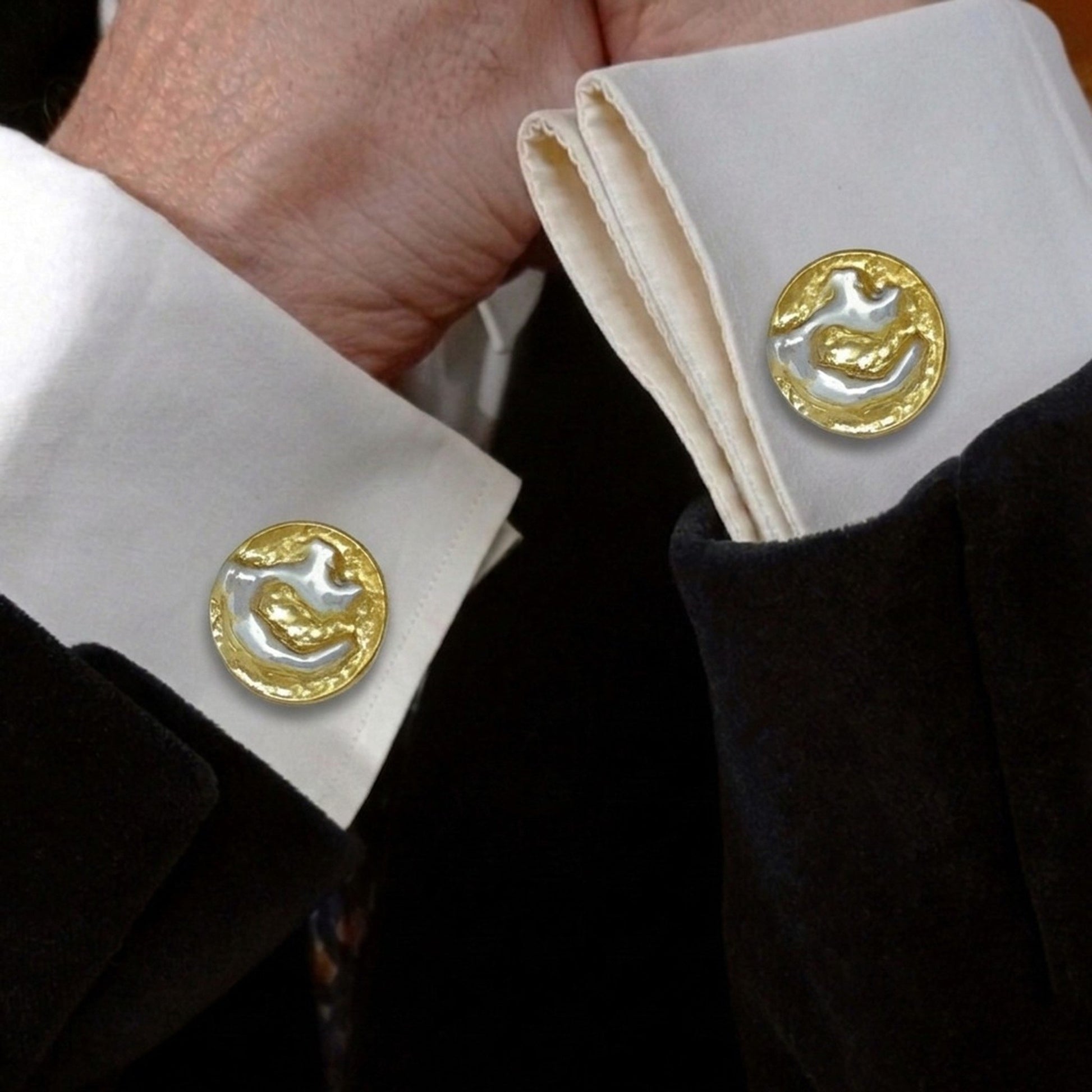 Detalle de los puños de una camisa blanca de vestir con unos gemelos circulares artesanales puestos. Los gemelos destacan por su relieve orgánico bicolor en plata y oro, contrastando con la manga de una chaqueta de traje oscura.