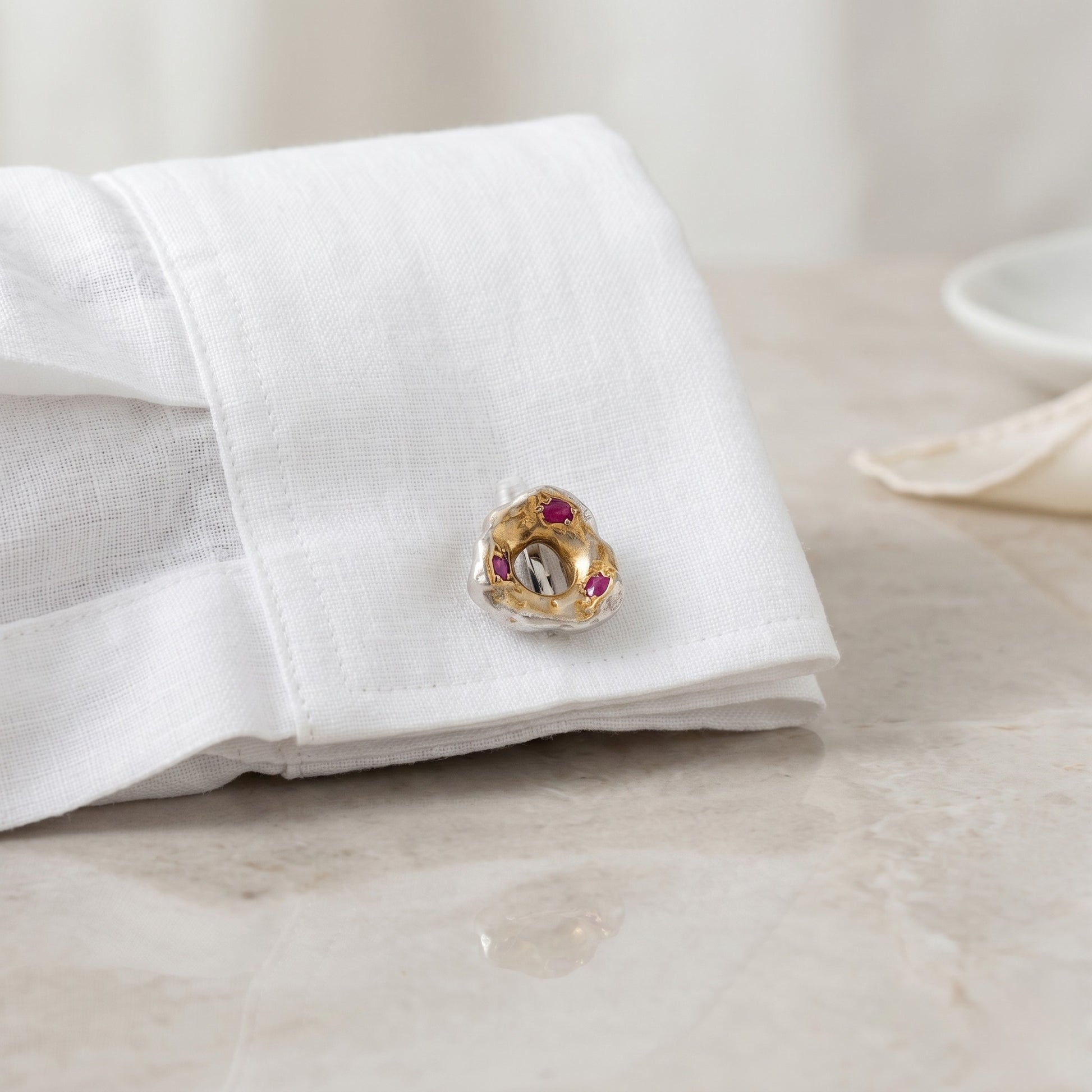 Detalle de gemelo de 1,4 cm de plata, oro y rubíes en el ojal de una camisa de lino blanca de alta calidad.