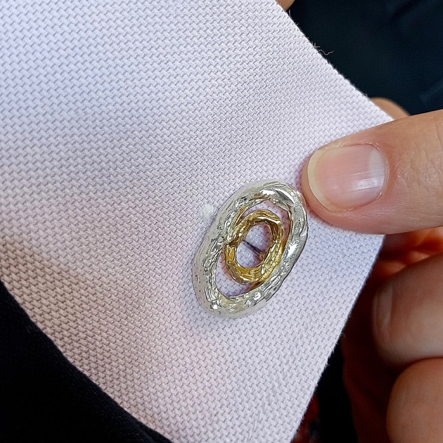 Un dedo índice sostiene un puño de camisa de tela texturizada de color rosa pálido, mostrando un gemelo de diseño 'Órbita' de plata texturizada y oro vermeil insertado en el ojal. El diseño elíptico se alinea con el puño.