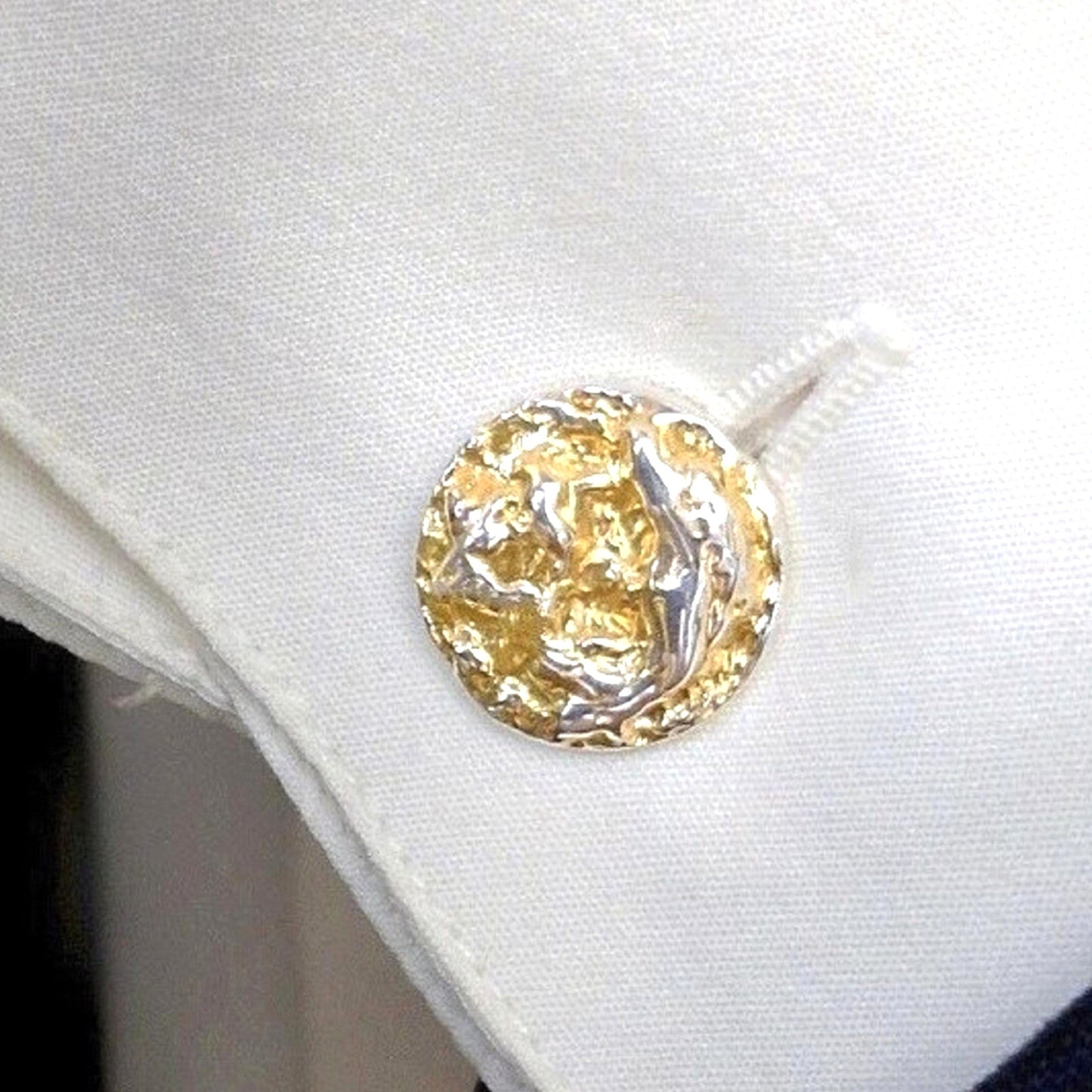 Una toma de primer plano que captura un solo gemelo circular con una cara de oro y plata texturizada a martillo. El gemelo está insertado de forma segura en el ojal de un puño de camisa de tela blanca nítida, que está perfectamente doblada.