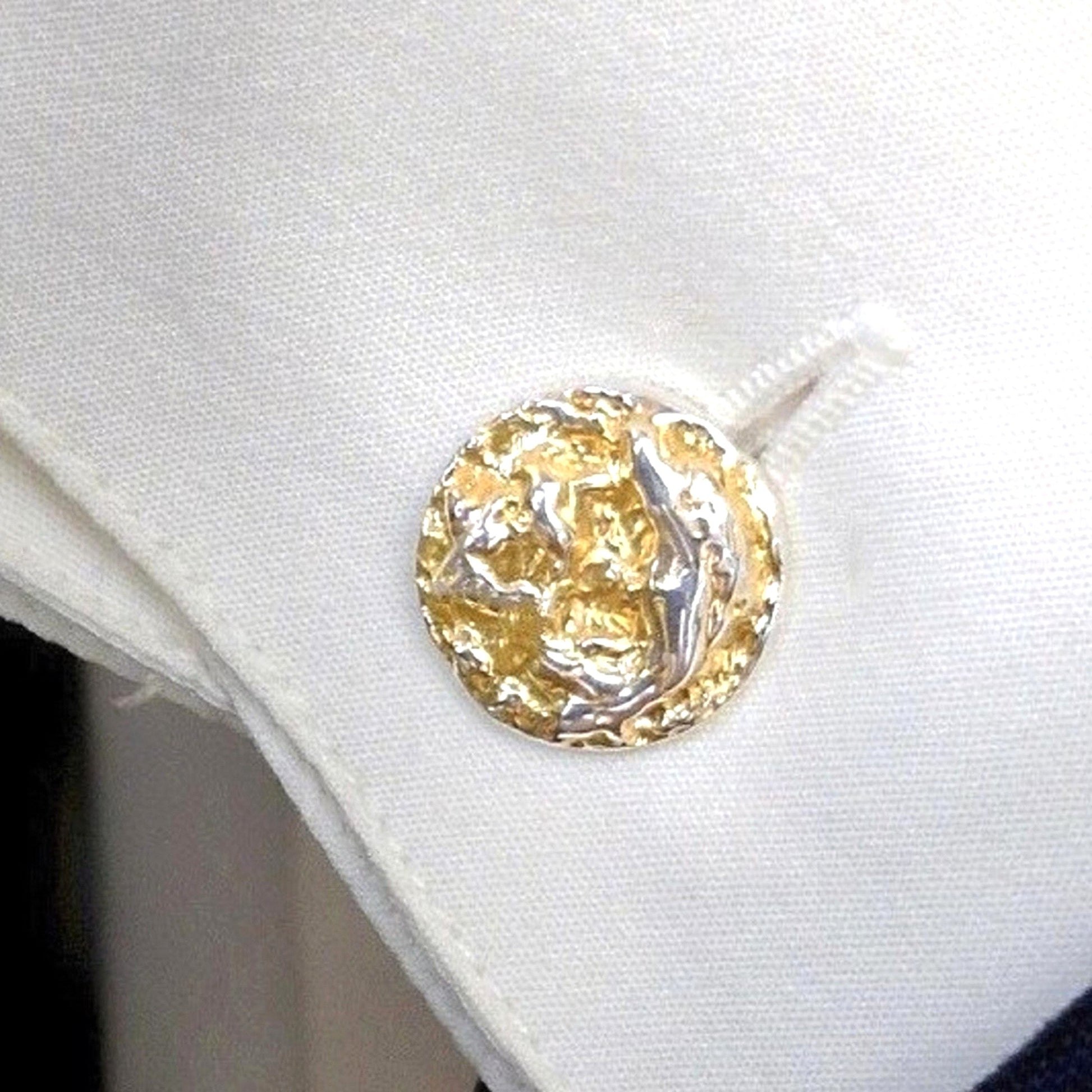 Una toma de primer plano que captura un solo gemelo circular con una cara de oro y plata texturizada a martillo. El gemelo está insertado de forma segura en el ojal de un puño de camisa de tela blanca nítida, que está perfectamente doblada.