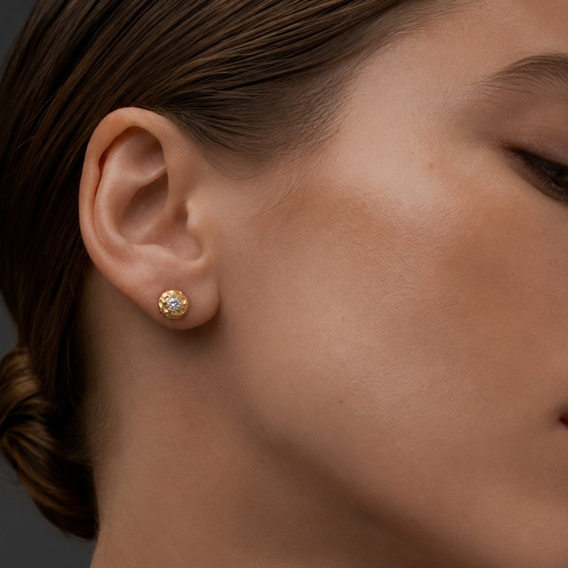 Vista de perfil de la MODELO ESPAÑOLA JOYERIA 1 luciendo el pendiente de botón redondo de oro texturizado con diamante en el lóbulo de la oreja con el cabello recogido.