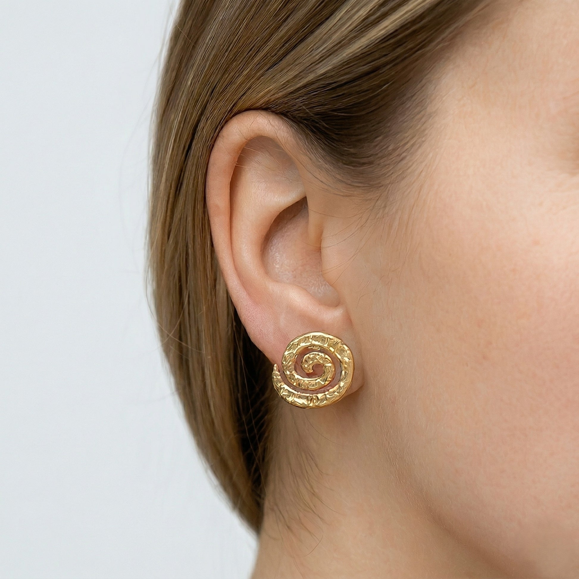 Primer plano detallado de la oreja de una mujer con cabello castaño, mostrando un pendiente de oro vermeil de 1,8 cm. El pendiente tiene un diseño de espiral martillada y orgánica, que se ajusta perfectamente al lóbulo. 