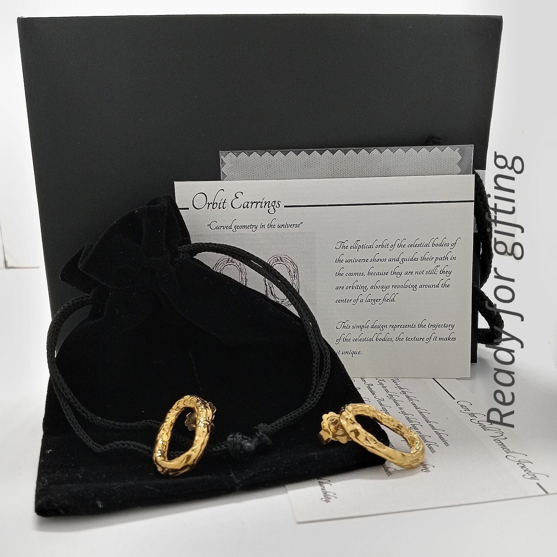 Un par de pendientes ovalados de oro vermeil texturizado sobre una bolsa de terciopelo negro. Detrás, una tarjeta de descripción "Orbit Earring" con texto explicativo, un paño de limpieza gris y una caja de regalo negra. A la derecha, un texto vertical que dice "Ready for gifting".