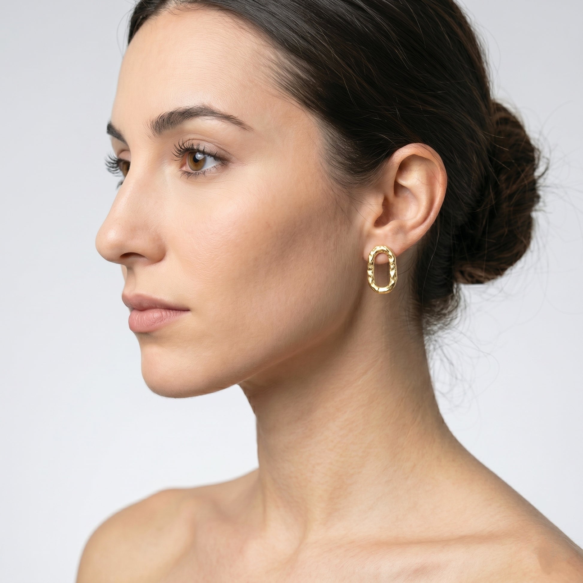 Primer plano de perfil de una mujer llevando un pendiente ovalado de oro vermeil texturizado (2,7x1,5 cm) en su oreja derecha. Su cabello está recogido en un moño. Fondo gris claro.