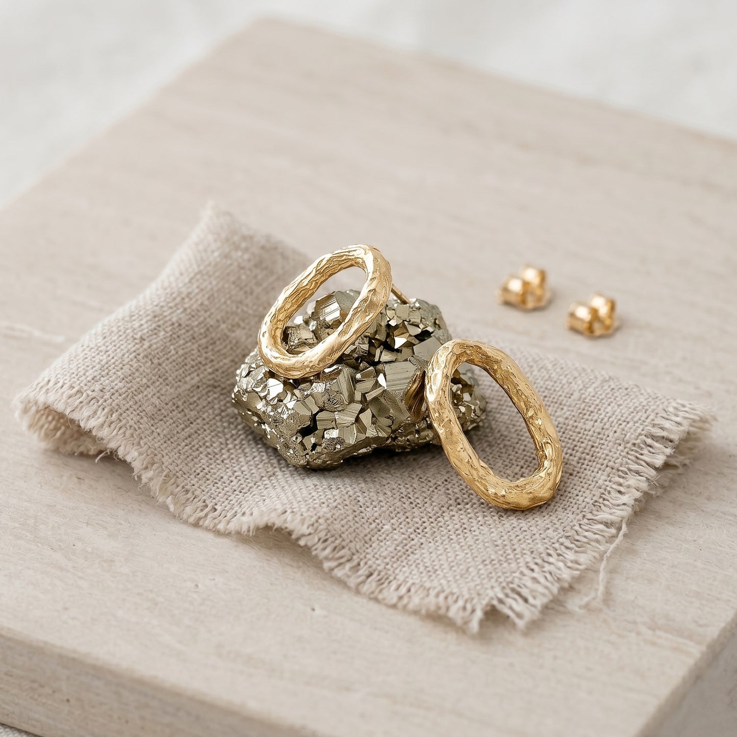 Un par de pendientes ovalados de oro vermeil con textura orgánica descansando sobre un trozo de mineral de pirita dorada, sobre una tela de lino texturizada y una base de madera clara. Se aprecian las dos presiones de cierre al fondo.