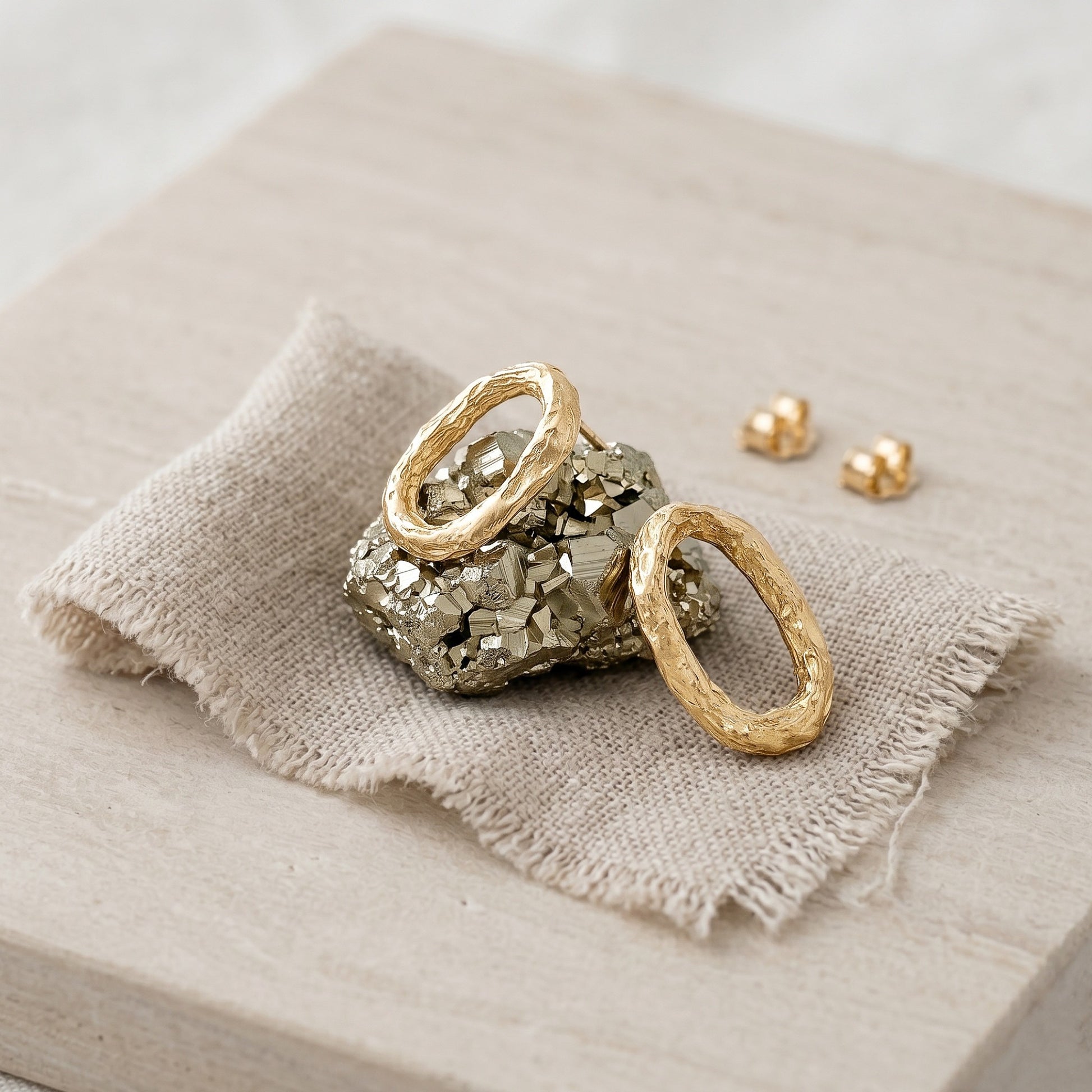 Un par de pendientes ovalados de oro vermeil con textura orgánica descansando sobre un trozo de mineral de pirita dorada, sobre una tela de lino texturizada y una base de madera clara. Se aprecian las dos presiones de cierre al fondo.