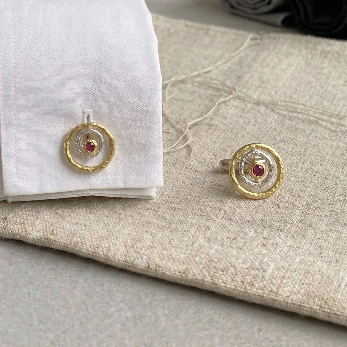 Gemelos artesanales de plata y oro vermeil con rubí rojo central, diseño circular inspirado en galaxias y formas cósmicas, colocados sobre puño de camisa blanca y tela de lino beige.