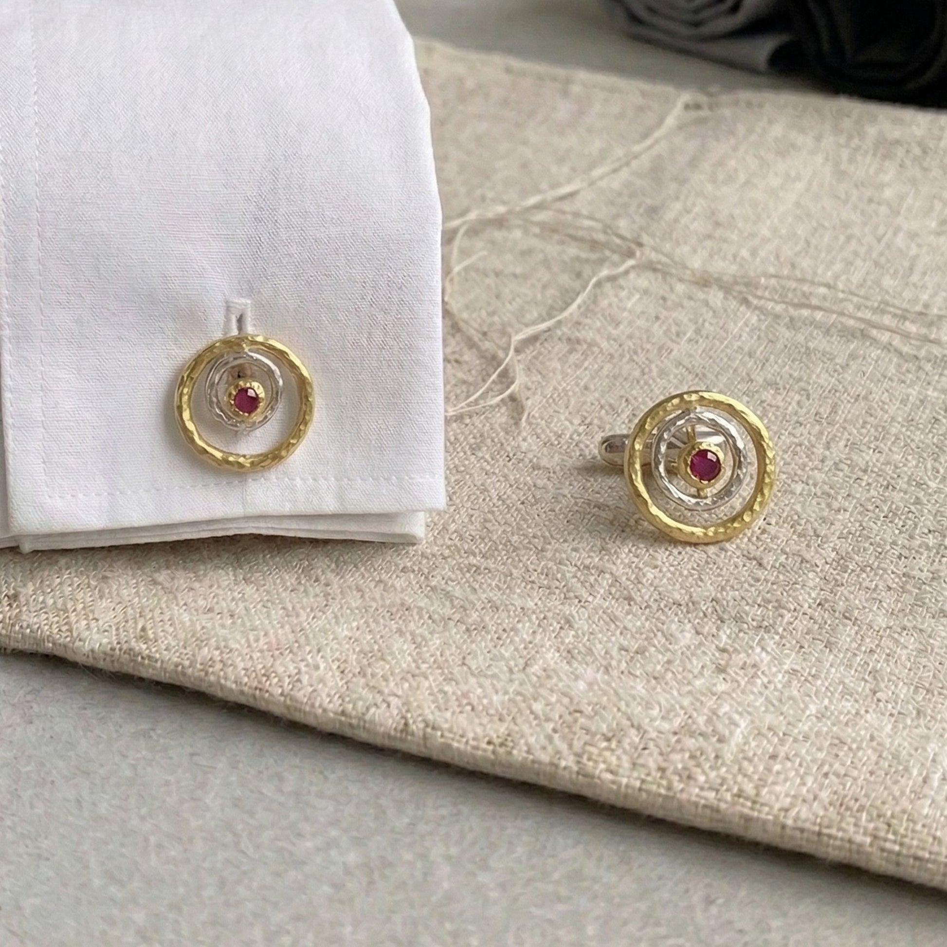 Gemelos artesanales de plata y oro vermeil con rubí rojo central, diseño circular inspirado en galaxias y formas cósmicas, colocados sobre puño de camisa blanca y tela de lino beige.
