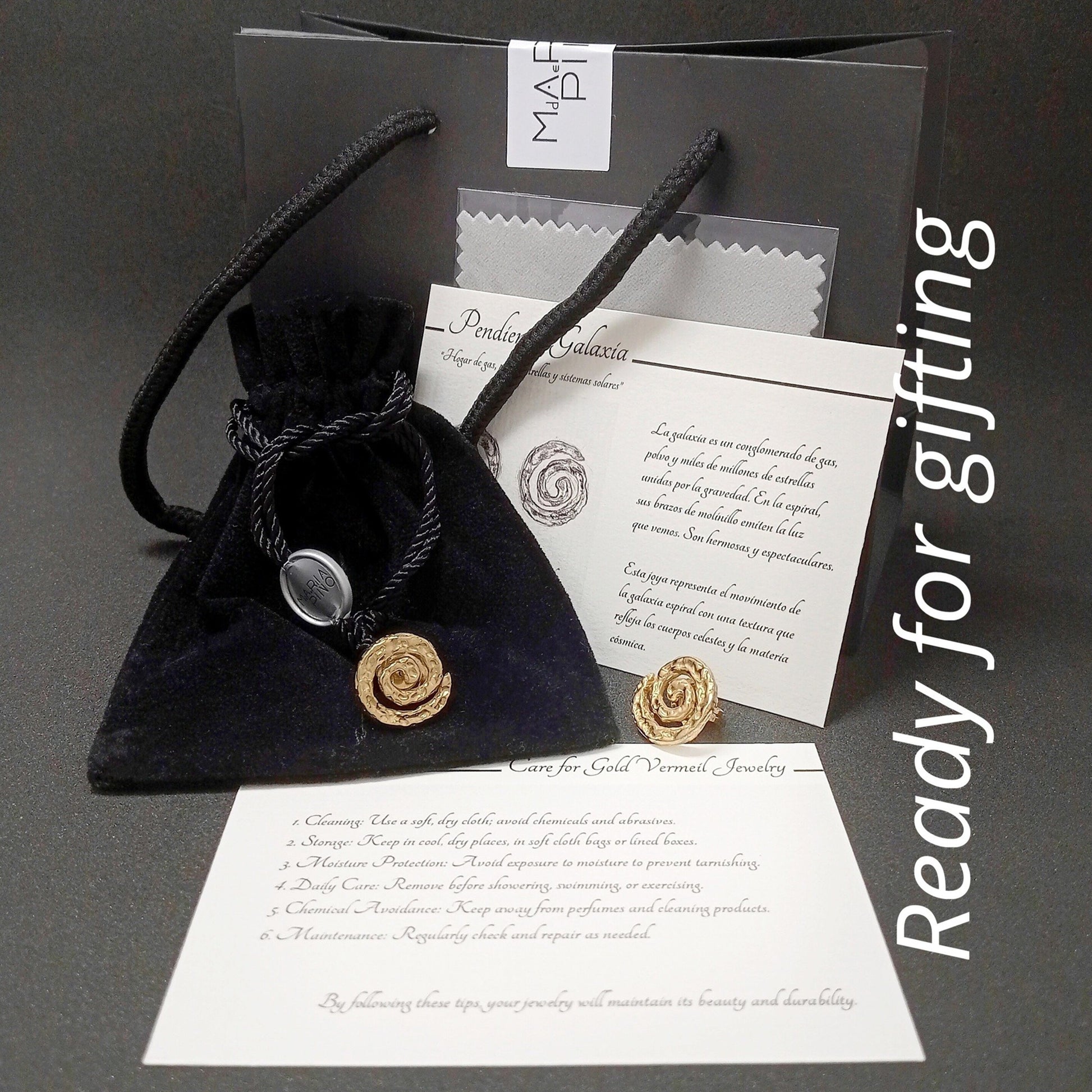 Kit de regalo de pendientes "Pendientes Galaxia" de oro vermeil de 1,8 cm. El set incluye: una bolsa de regalo de papel negra, una bolsa de terciopelo negra, un paño de limpieza, un par de pendientes de oro martillado en forma de espiral y dos tarjetas informativas. Una tarjeta detalla la inspiración cósmica y la otra los cuidados de la joyería.